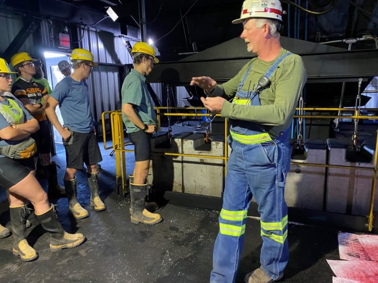 A man in a hard hat and blue overalls speaks to a group of people in hard hats and rubber boots inside an industrial facility, preparing for an expedition like those in Glacier National Park, surrounded by metal railings and machinery.