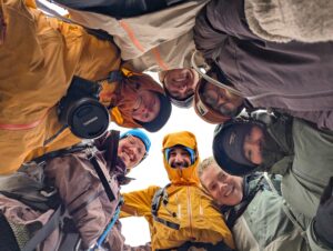 Six people in outdoor gear stand in a tight circle, looking down at the camera and smiling. The sky is bright above them, and they appear to be on an outdoor adventure in cold weather.