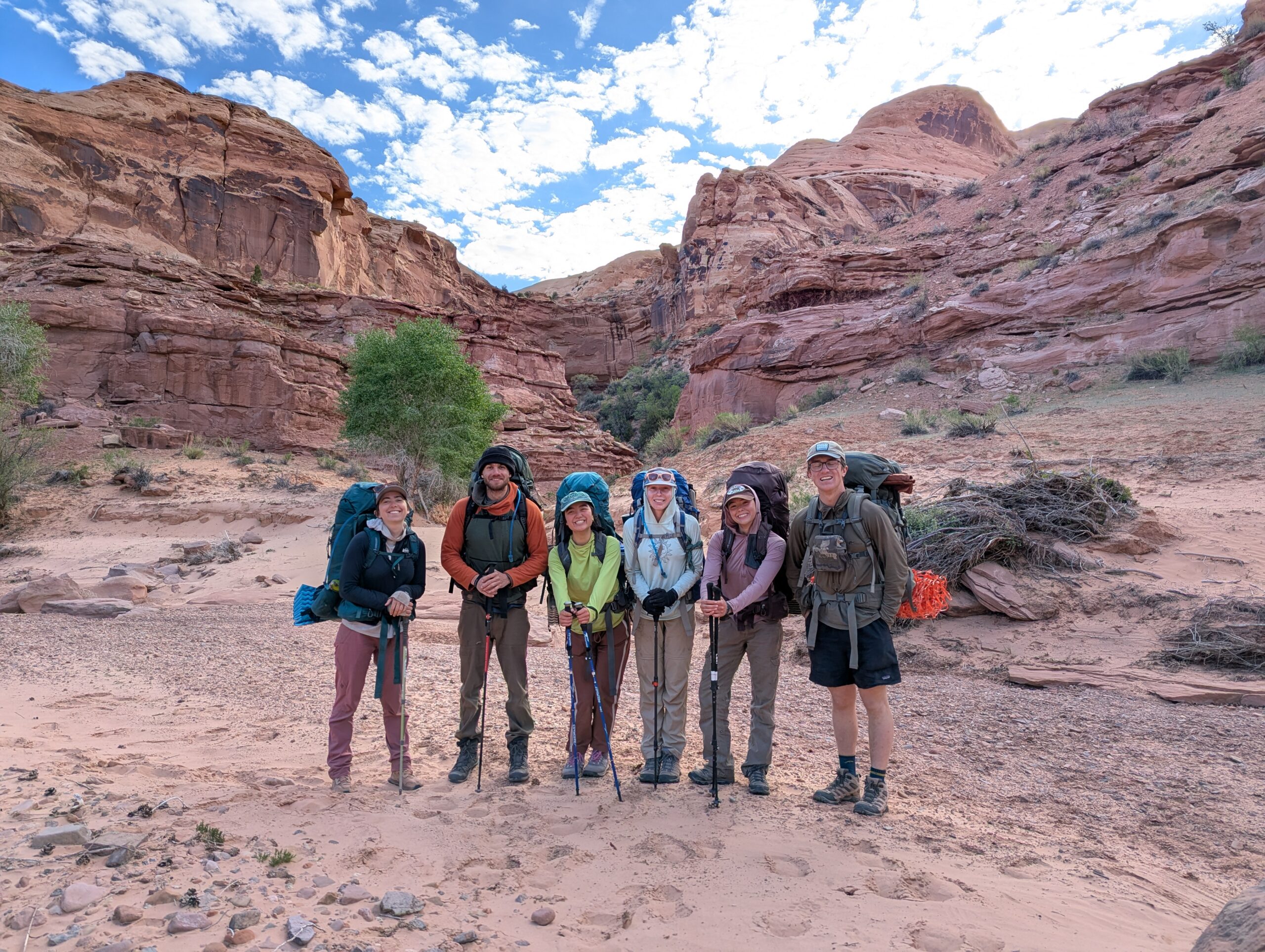 Six hikers with backpacks and trekking poles stand smiling in a rocky desert canyon with red rock formations, sparse vegetation, and a partly cloudy sky in the background.