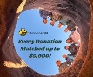 A group of people smile and look down into a rock formations opening. Text reads: Missoula Gives. Every Donation Matched up to $5,000! against a blue sky background.