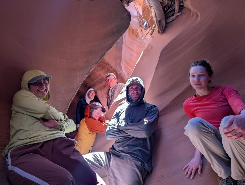 Six people sit and pose inside a narrow, sunlit slot canyon with smooth, curving sandstone walls. Some wear hats and hoodies, and light streams through an opening above them.