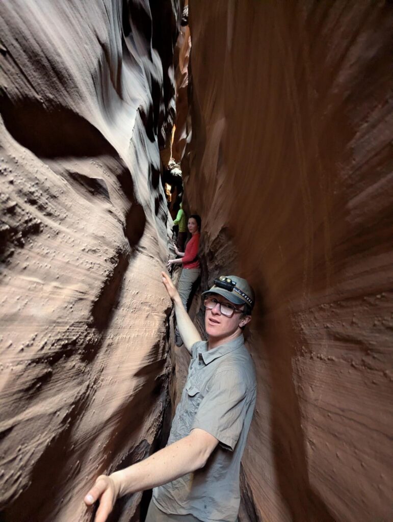 Two people carefully make their way through a narrow slot canyon with smooth, towering rock walls close on either side. The person in front wears glasses and a hat, while the other is further back in the canyon.