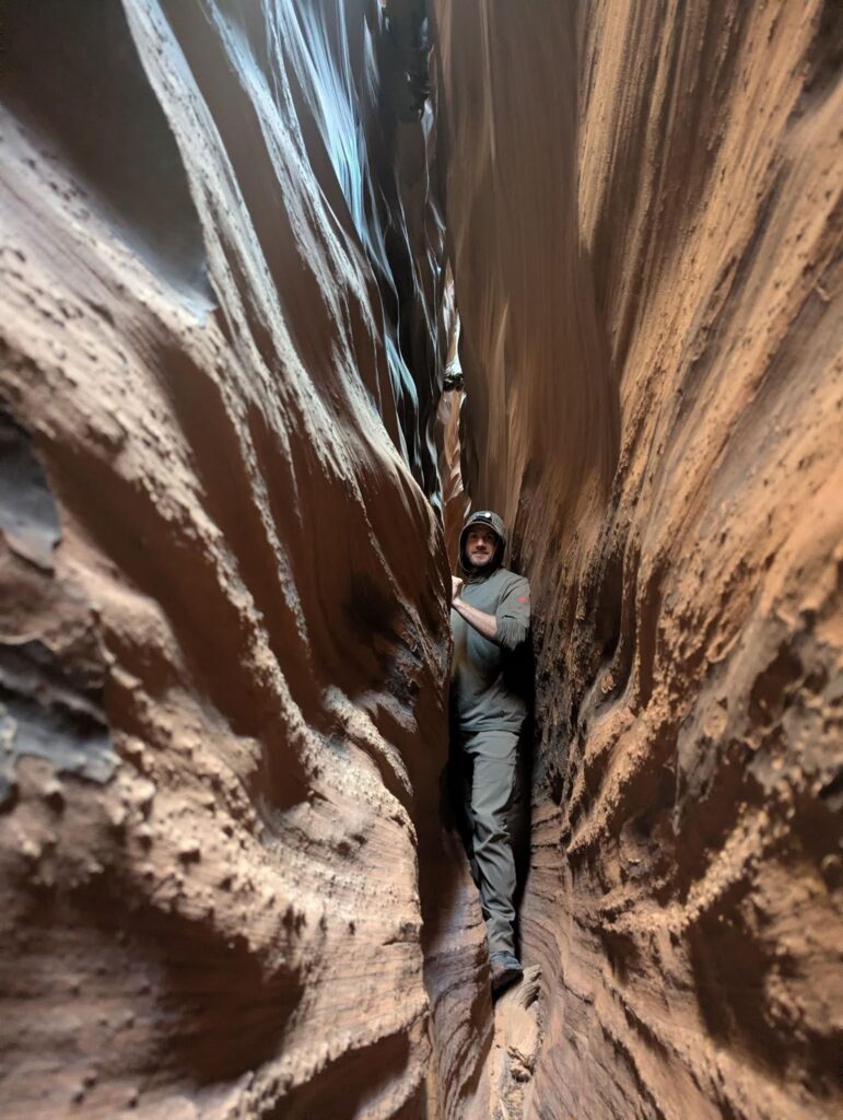 A person wearing outdoor clothing stands sideways in a narrow, winding slot canyon with tall, textured rock walls close on both sides. Light filters down from above, illuminating the earthy tones of the canyon.