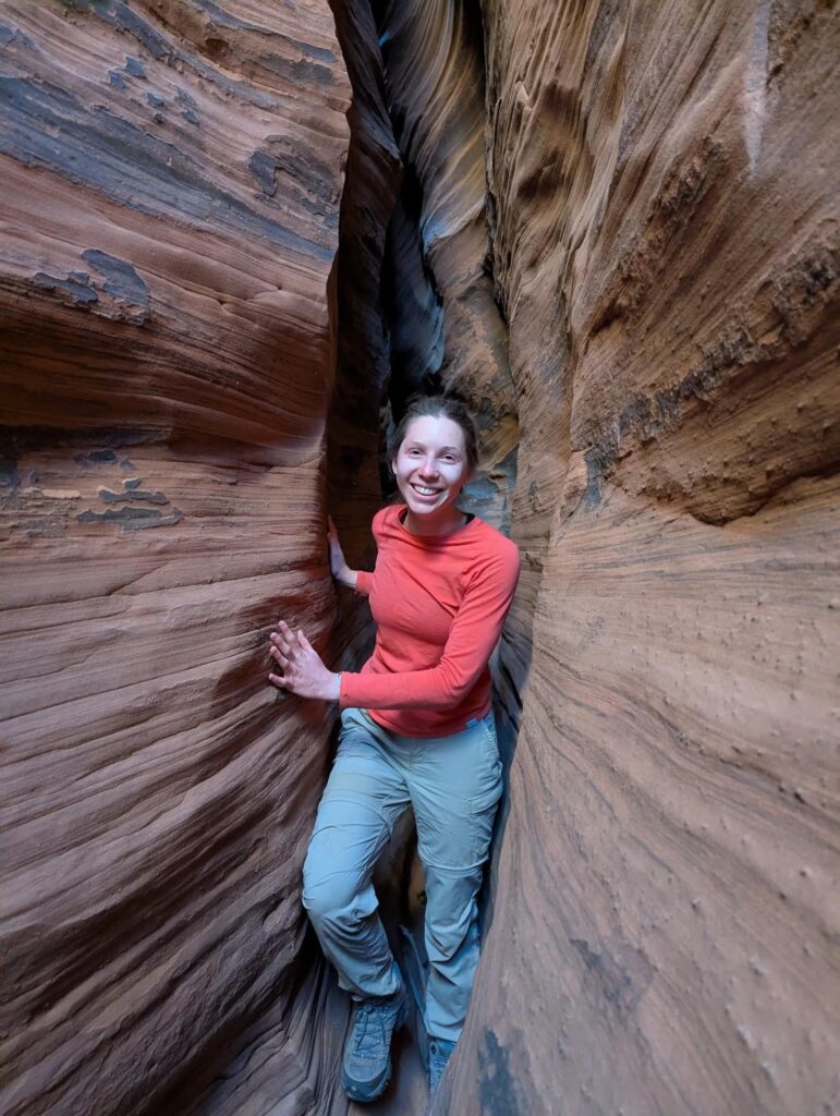 A woman in an orange long-sleeve shirt and light pants stands smiling in a narrow slot canyon with layered, textured rock walls close on either side.