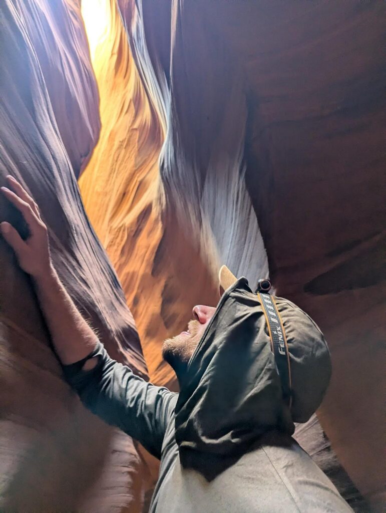 A person wearing a hooded jacket looks up while standing between the narrow, winding sandstone walls of a slot canyon, illuminated by sunlight streaming from above.